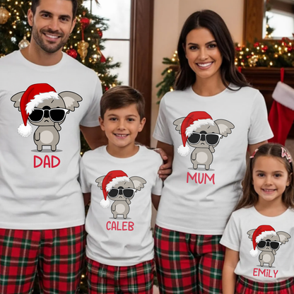A family of four wearing matching white graphic t-shirts and red plaid pajama pants, standing together in a decorated Christmas living room. Each person's shirt features a cartoon gray angel with sunglasses and a red Santa hat, with 'DAD', 'MUM', 'CALEB', and 'EMILY' printed in red below.