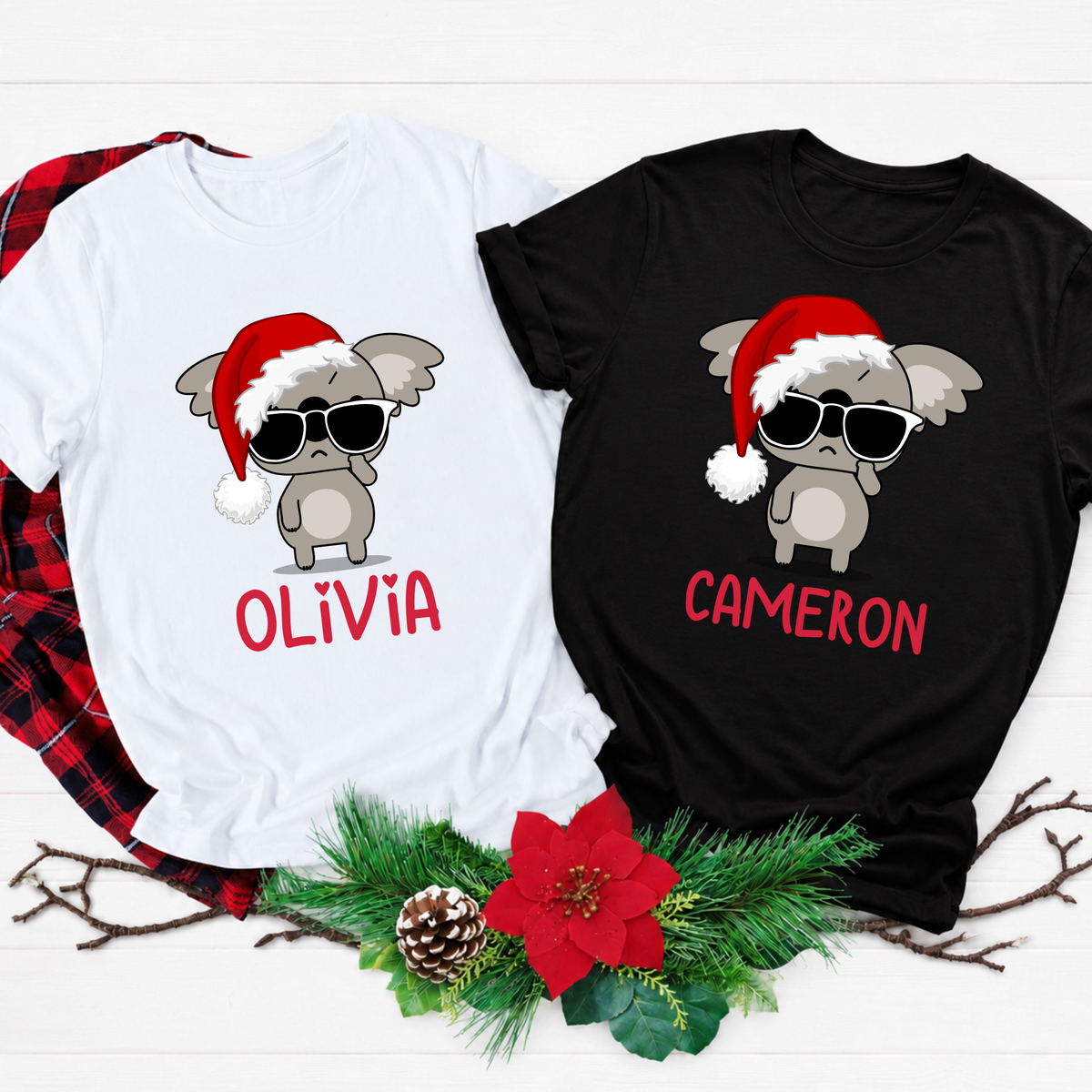 Two custom Christmas graphic t-shirts, one white and one black, displayed on a white wood background with a red plaid shirt underneath and holiday decorations at the bottom. The shirts feature a cartoon gray baby koala wearing a red Santa hat and sunglasses, with the names 'OLIVIA' and 'CAMERON' printed in red below.