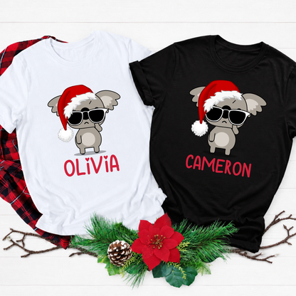 Two custom Christmas graphic t-shirts, one white and one black, displayed on a white wood background with a red plaid shirt underneath and holiday decorations at the bottom. The shirts feature a cartoon gray baby koala wearing a red Santa hat and sunglasses, with the names 'OLIVIA' and 'CAMERON' printed in red below.