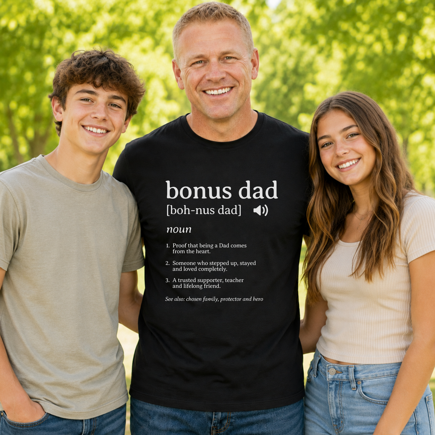 Three people standing outdoors in front of a blurred green background, with the center person wearing a black graphic T-shirt that reads 'bonus dad [boh-nus dad]' with a dictionary-style definition. Because people and a real outdoor environment are visible, this is Lifestyle rather than Product/Packaging.