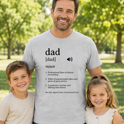 A man and two children standing together in a sunny park, with the man wearing a light gray graphic T-shirt featuring a dictionary-style definition of 'dad'.