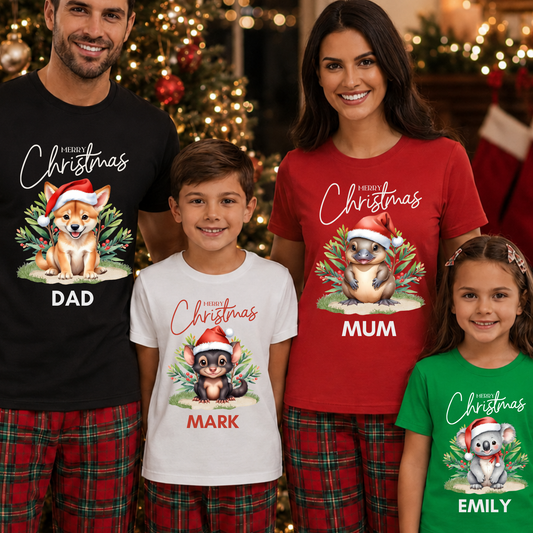 A family of four wearing matching Christmas graphic t-shirts and plaid pajama pants in front of a decorated Christmas tree. The shirts feature different animal illustrations (a corgi, a beaver, a koala) with Santa hats, and the text 'Merry Christmas' and the names 'DAD', 'MUM', 'MARK', and 'EMILY'.