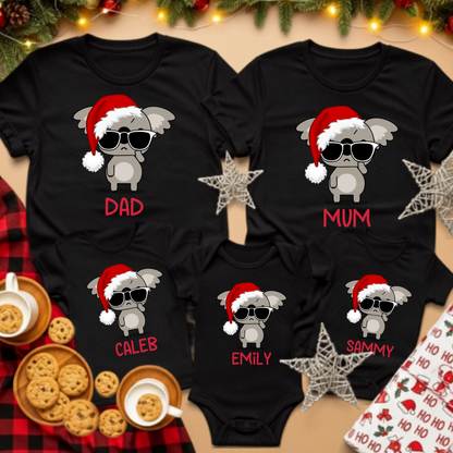Five black baby bodysuits featuring a matching Christmas graphic of a gray cartoon elephant wearing a Santa hat and sunglasses, with personalized names printed in red below each design. The shirts are arranged in a flat-lay holiday setting with cookies, coffee cups, pine branches, and gift wrapping.