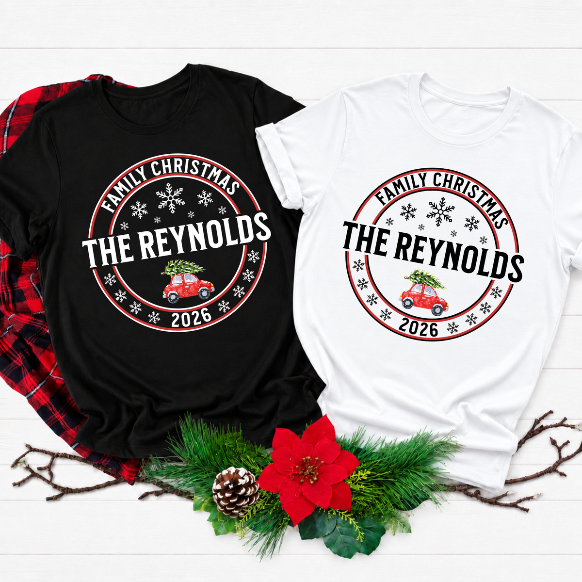 Two custom Christmas t-shirts displayed on a white wood background with holiday greenery and a red plaid shirt prop. One shirt is black and the other is white, both featuring a circular graphic that reads 'Family Christmas The Reynolds 2026' with a red vintage car carrying a Christmas tree.