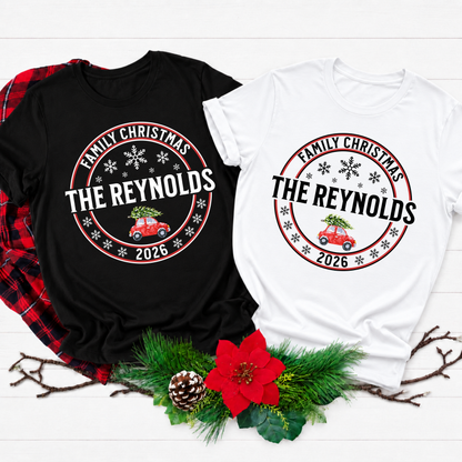 Two custom Christmas t-shirts displayed on a white wood background with holiday greenery and a red plaid shirt prop. One shirt is black and the other is white, both featuring a circular graphic that reads 'Family Christmas The Reynolds 2026' with a red vintage car carrying a Christmas tree.