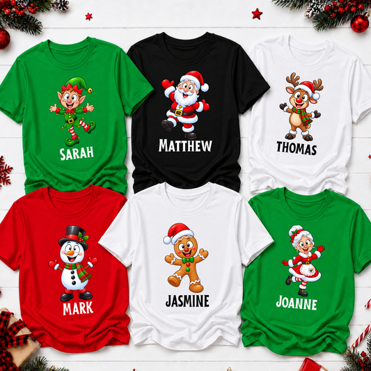 Six personalized Christmas graphic t-shirts in green, black, white, and red, arranged in a flat-lay on a white wood background with holiday decorations. The shirts feature cartoon holiday characters (elf, Santa, reindeer, snowman, gingerbread man, and Santa with a child) and are personalized with the names Sarah, Matthew, Thomas, Mark, Jasmine, and Joanne printed below the graphics.