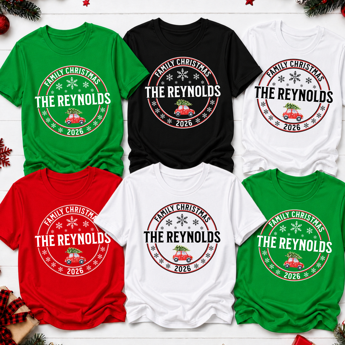 Six Christmas-themed t-shirts in green, black, and white, arranged in a 2x3 grid on a white wood background. Each shirt features a circular graphic with the text 'FAMILY CHRISTMAS THE REYNOLDS 2026' and a small red truck carrying a green tree, decorated with snowflakes.