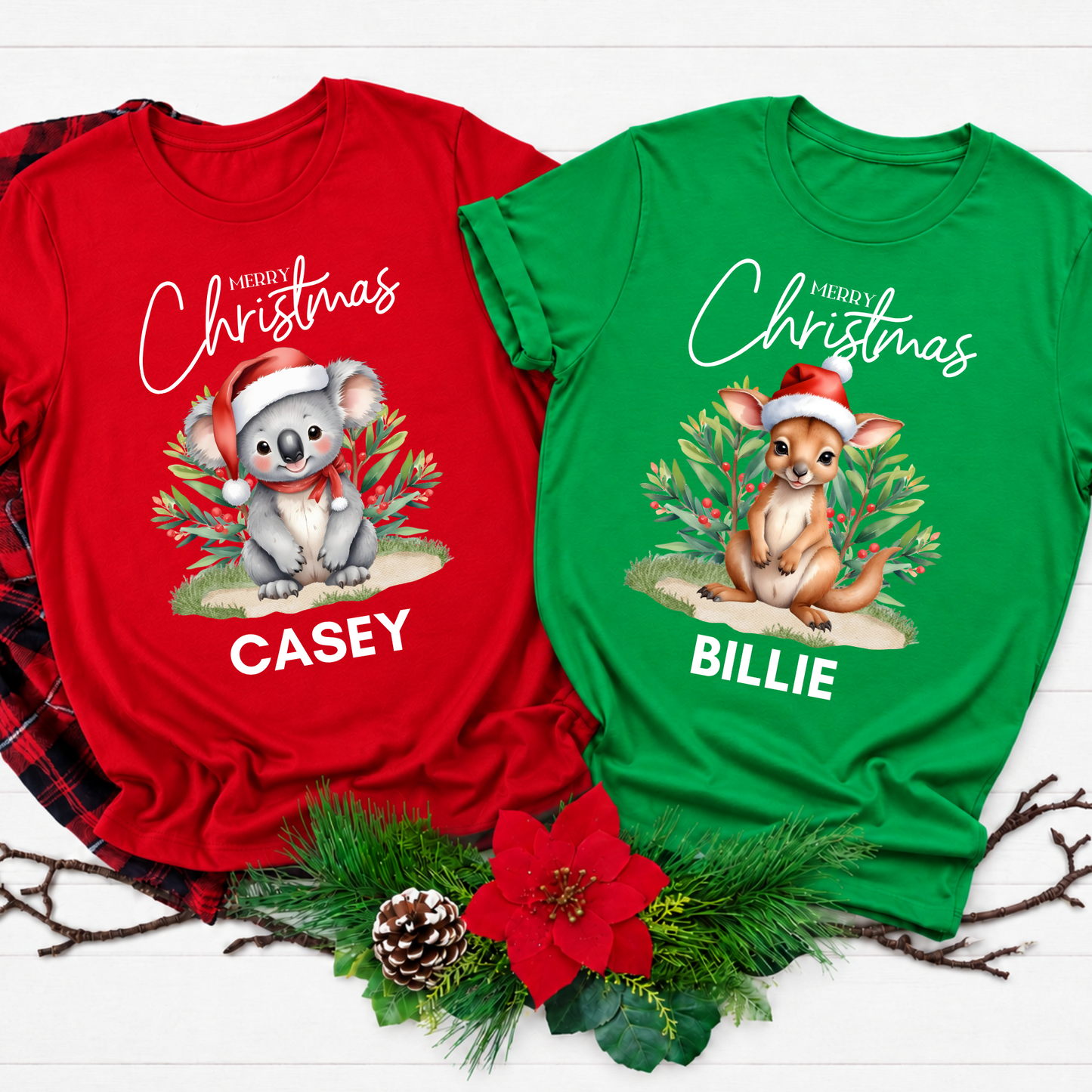 Two personalized Christmas graphic t-shirts displayed on a white wood background with holiday decorations. The left shirt is red and features a koala wearing a Santa hat with the text 'Merry Christmas Casey'.