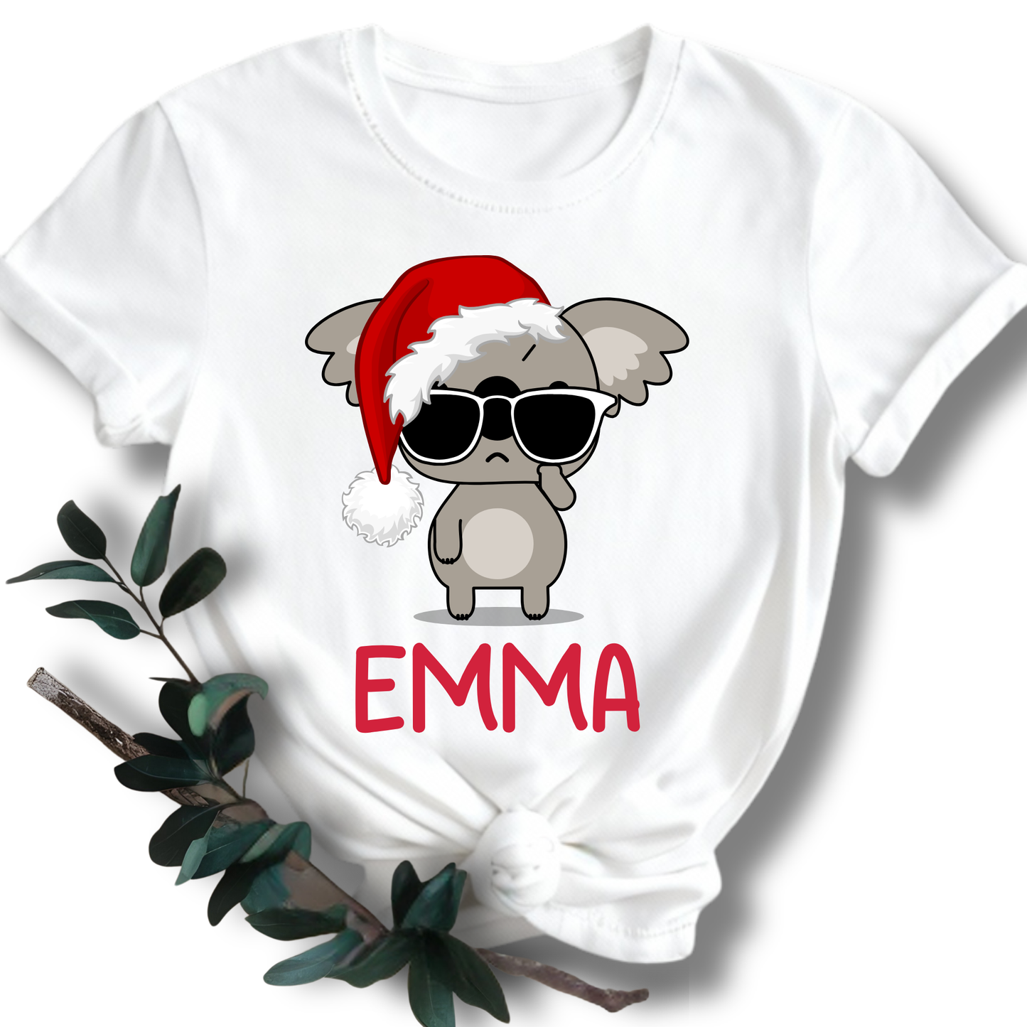 A white short-sleeve T-shirt with a graphic of a gray cartoon koala wearing a red Santa hat and black sunglasses, with the name 'EMMA' printed in red below. The shirt is styled flat with a knot at the bottom and green leaves on a white background.