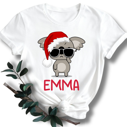 A white short-sleeve T-shirt with a graphic of a gray cartoon koala wearing a red Santa hat and black sunglasses, with the name 'EMMA' printed in red below. The shirt is styled flat with a knot at the bottom and green leaves on a white background.