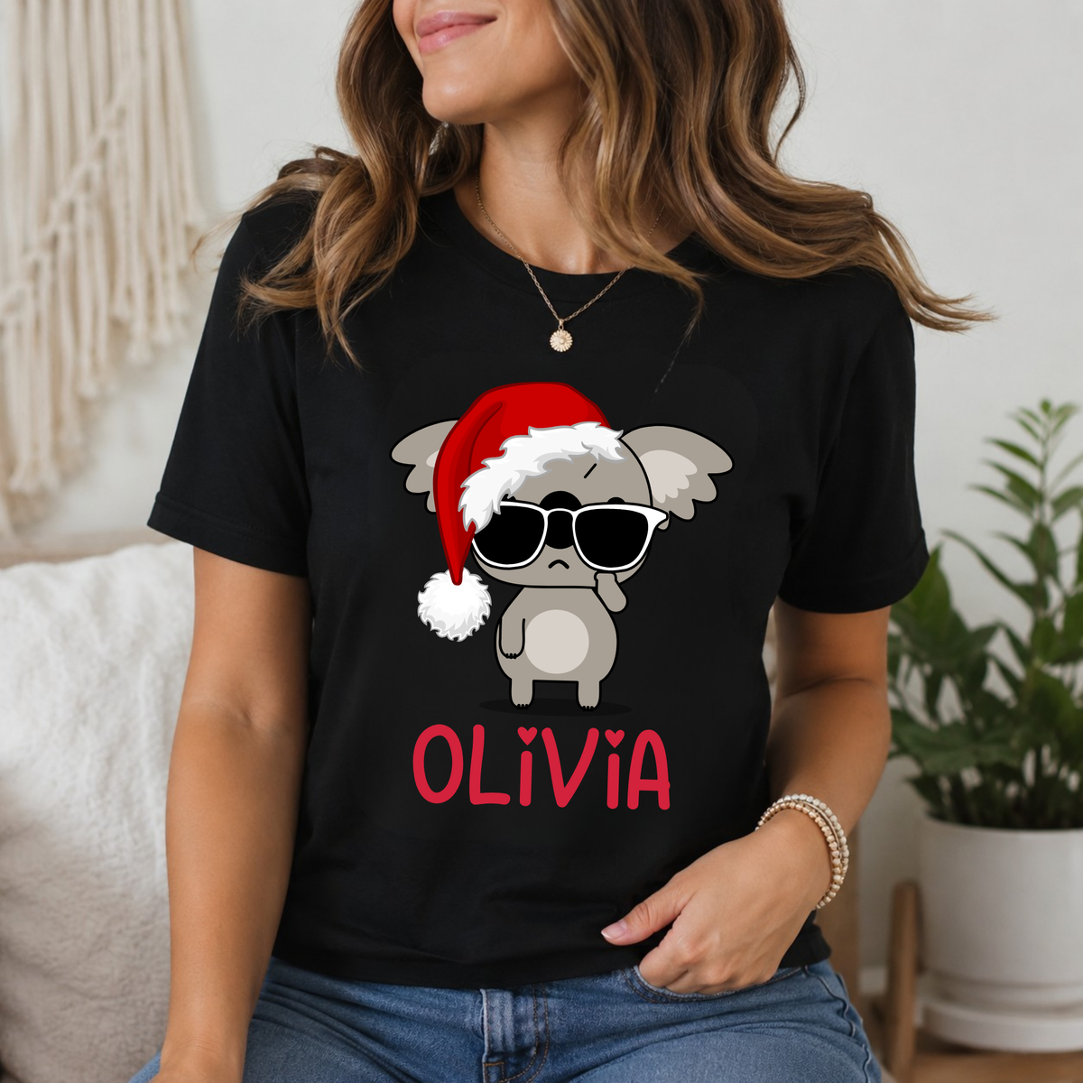 A woman wearing a black graphic t-shirt with a cartoon gray creature wearing a red Santa hat and sunglasses, with the name 'OLIVIA' printed in red below. The photo is taken in a cozy indoor setting with a plant in the background.