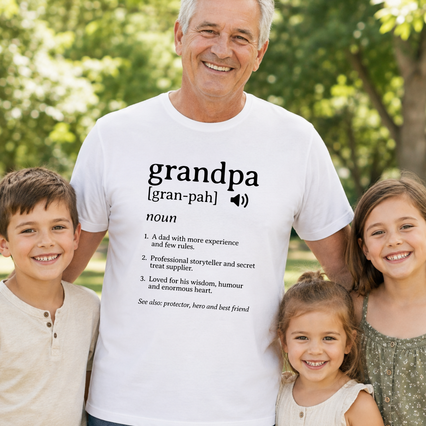 A smiling older man wearing a white graphic T-shirt stands outdoors with three children. The shirt has a dictionary-style definition printed in black text reading 'grandpa [gran-pah] noun' with a pronunciation icon and a list of meanings.